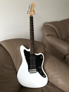 A white electric guitar with a black pickguard is resting on a beige leather couch. The guitar has a maple neck and headstock with chrome tuning pegs and features a classic design.