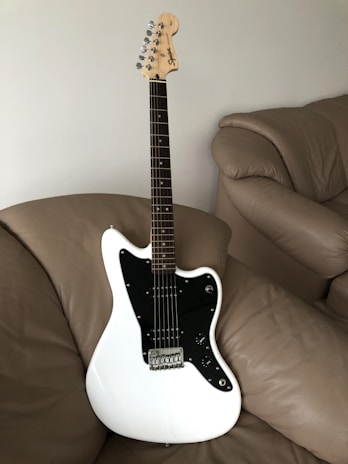 A white electric guitar with a black pickguard is resting on a beige leather couch. The guitar has a maple neck and headstock with chrome tuning pegs and features a classic design.