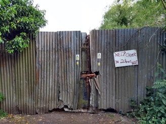 A rusty corrugated metal gate is flanked by lush green foliage on both sides. The gate, showing signs of wear and tear, has a visible gap in the middle and a handwritten sign that reads 'KEEP clear IN 24hr USE' attached to it. Warning stickers are also visible on the gate.