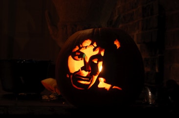 A carved pumpkin features an intricate face design illuminated from within by a light source, creating a glowing effect against the dark background.
