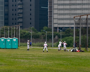 A group of people wearing baseball uniforms are practicing on a grassy field. There are several players spread across the field, with one appearing to be preparing to pitch while others are positioned in the fielding formation. Three portable toilets are visible to the left, and city buildings loom in the background, indicating an urban setting.