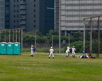 A group of people wearing baseball uniforms are practicing on a grassy field. There are several players spread across the field, with one appearing to be preparing to pitch while others are positioned in the fielding formation. Three portable toilets are visible to the left, and city buildings loom in the background, indicating an urban setting.