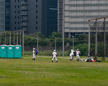 A group of people wearing baseball uniforms are practicing on a grassy field. There are several players spread across the field, with one appearing to be preparing to pitch while others are positioned in the fielding formation. Three portable toilets are visible to the left, and city buildings loom in the background, indicating an urban setting.