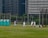 A group of people wearing baseball uniforms are practicing on a grassy field. There are several players spread across the field, with one appearing to be preparing to pitch while others are positioned in the fielding formation. Three portable toilets are visible to the left, and city buildings loom in the background, indicating an urban setting.