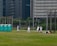 A group of people wearing baseball uniforms are practicing on a grassy field. There are several players spread across the field, with one appearing to be preparing to pitch while others are positioned in the fielding formation. Three portable toilets are visible to the left, and city buildings loom in the background, indicating an urban setting.