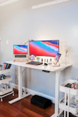 A modern home office setup with a white standing desk featuring a large monitor, a laptop, and a smartphone, all displaying a colorful screensaver. The desk also holds a keyboard, mouse, small plants, and a lamp. Nearby, there are storage carts containing books and office supplies. The room has a light grey wall and wooden flooring.