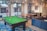 A modern indoor recreational room featuring a green pool table with balls arranged on the table, located on a patterned blue rug. Adjacent to the pool table is a ping pong table with a net. The room includes a comfortable blue sectional sofa with colorful cushions. Large windows allow natural light to illuminate the space, offering a view of an outdoor area with garage doors. The room also features pendant lighting and a small kitchenette area with a stainless steel refrigerator and decorative wall art.