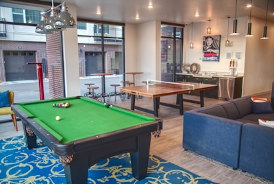 A modern indoor recreational room featuring a green pool table with balls arranged on the table, located on a patterned blue rug. Adjacent to the pool table is a ping pong table with a net. The room includes a comfortable blue sectional sofa with colorful cushions. Large windows allow natural light to illuminate the space, offering a view of an outdoor area with garage doors. The room also features pendant lighting and a small kitchenette area with a stainless steel refrigerator and decorative wall art.