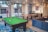 A modern indoor recreational room featuring a green pool table with balls arranged on the table, located on a patterned blue rug. Adjacent to the pool table is a ping pong table with a net. The room includes a comfortable blue sectional sofa with colorful cushions. Large windows allow natural light to illuminate the space, offering a view of an outdoor area with garage doors. The room also features pendant lighting and a small kitchenette area with a stainless steel refrigerator and decorative wall art.