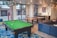 A modern indoor recreational room featuring a green pool table with balls arranged on the table, located on a patterned blue rug. Adjacent to the pool table is a ping pong table with a net. The room includes a comfortable blue sectional sofa with colorful cushions. Large windows allow natural light to illuminate the space, offering a view of an outdoor area with garage doors. The room also features pendant lighting and a small kitchenette area with a stainless steel refrigerator and decorative wall art.
