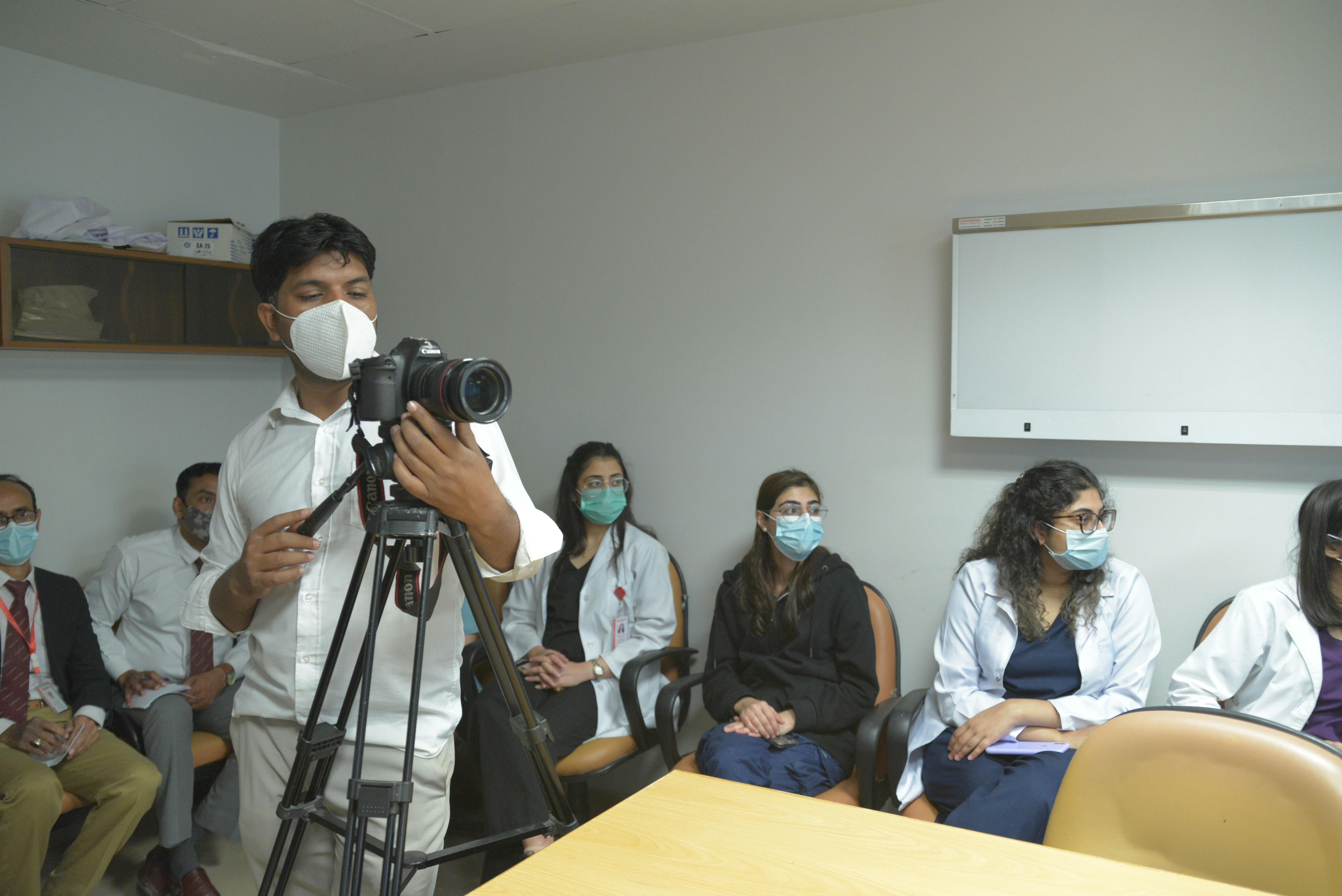 A group of people are gathered in a clinical or educational setting. Several individuals are seated, wearing masks and professional attire, including white coats. One person is standing in the foreground operating a camera mounted on a tripod, also wearing a mask.