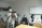 A group of people are gathered in a clinical or educational setting. Several individuals are seated, wearing masks and professional attire, including white coats. One person is standing in the foreground operating a camera mounted on a tripod, also wearing a mask.