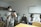 A group of people are gathered in a clinical or educational setting. Several individuals are seated, wearing masks and professional attire, including white coats. One person is standing in the foreground operating a camera mounted on a tripod, also wearing a mask.