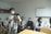 A group of people are gathered in a clinical or educational setting. Several individuals are seated, wearing masks and professional attire, including white coats. One person is standing in the foreground operating a camera mounted on a tripod, also wearing a mask.