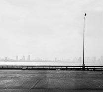 A black-and-white image featuring an expanse of road in the foreground, with a single streetlight standing tall on the right side. The background shows a hazy skyline of distant buildings, possibly indicating a cityscape shrouded in fog or mist. The scene exudes an atmospheric and minimalistic quality.