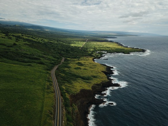 A scenic coastal landscape featuring a winding road that runs alongside lush green fields and hills. The coastline meets the sea with gentle waves lapping against the rocky shoreline. The sky is partially cloudy, casting soft shadows over the land and sea.