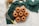 A group of seven small bundt cakes is arranged on a circular wire rack. The cakes are golden-brown and set against a textured green fabric backdrop. Nearby, there are two spoons, a coil of twine, some sprigs of herbs, and a whisk with a yellow handle.
