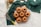 A group of seven small bundt cakes is arranged on a circular wire rack. The cakes are golden-brown and set against a textured green fabric backdrop. Nearby, there are two spoons, a coil of twine, some sprigs of herbs, and a whisk with a yellow handle.