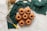 A group of seven small bundt cakes is arranged on a circular wire rack. The cakes are golden-brown and set against a textured green fabric backdrop. Nearby, there are two spoons, a coil of twine, some sprigs of herbs, and a whisk with a yellow handle.