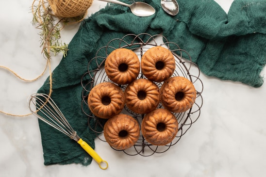 A group of seven small bundt cakes is arranged on a circular wire rack. The cakes are golden-brown and set against a textured green fabric backdrop. Nearby, there are two spoons, a coil of twine, some sprigs of herbs, and a whisk with a yellow handle.
