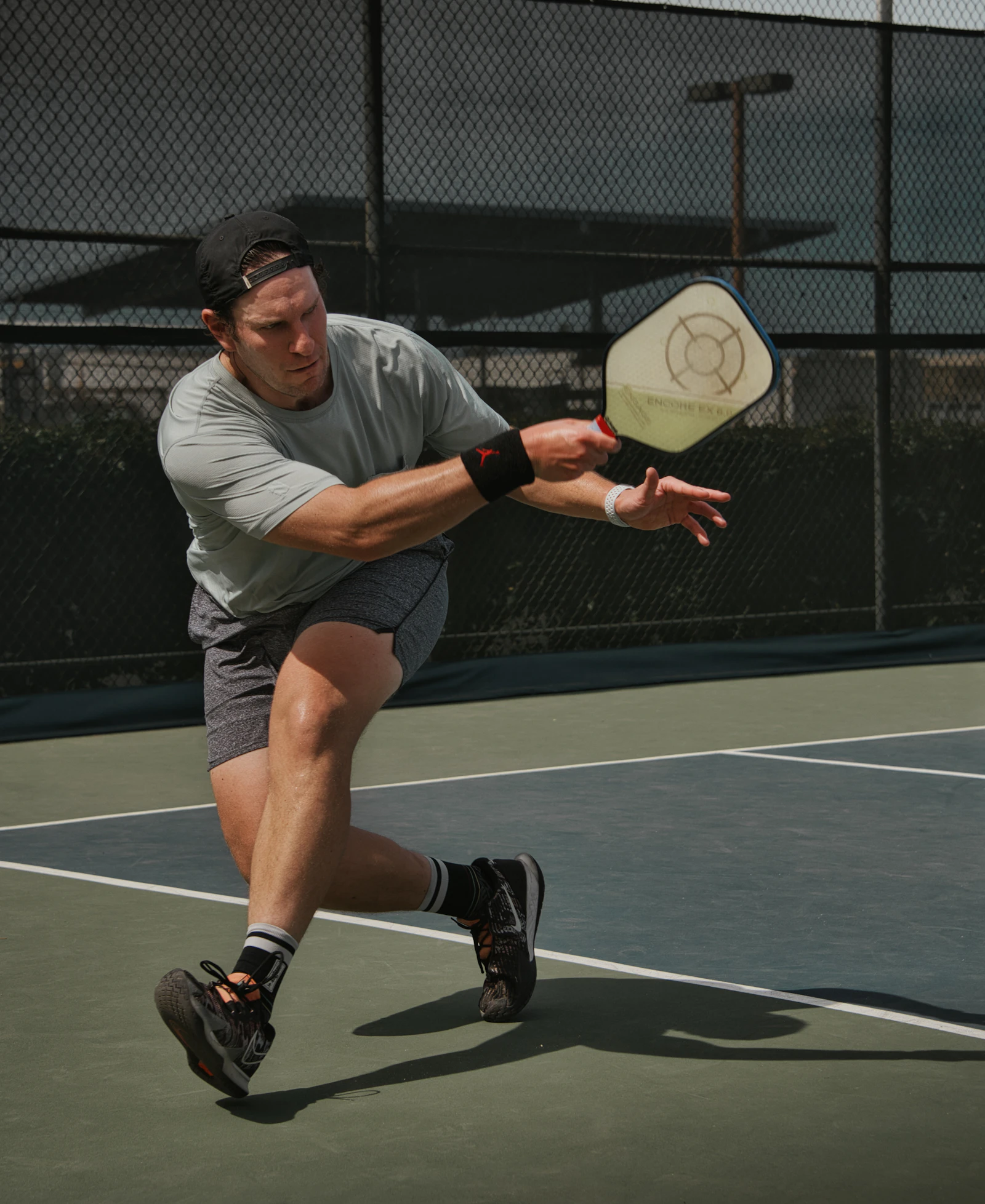A man lunges to return a shot with a solid faced pickleball paddle on a blue and green court behind a chain link fence.