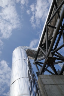 A large metallic structure with reflective surfaces extends upward into the sky, where soft, scattered clouds are visible. The structure includes an industrial-looking pipe or duct system and a sturdy, angular metal frame supported by a concrete base.