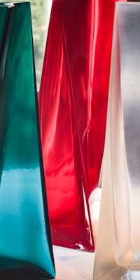 Glossy and translucent vase-like structures with smooth surfaces in rich colors, including teal, red, and a silvery hue. These forms stand close together, showcasing a sleek modern design.