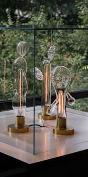 Three artistically crafted glass light bulbs with unique and abstract shapes are displayed within a glass case. They are set on brass bases and emit warm, orange filament glows. The background features blurred green foliage, providing a natural contrast to the modern aesthetic of the bulbs.