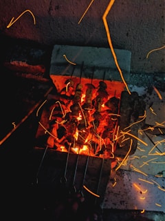 Skewers of food are being grilled on a barbecue with glowing coals. Sparks and embers are visible, flying in different directions, creating a vibrant and dynamic effect. The setup appears to be in a dimly lit outdoor area.
