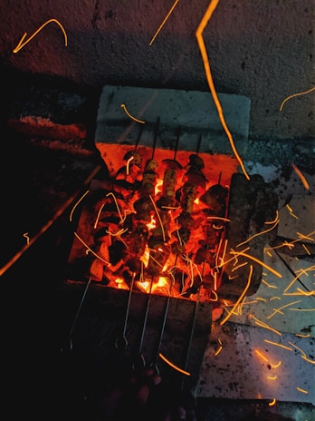 Skewers of food are being grilled on a barbecue with glowing coals. Sparks and embers are visible, flying in different directions, creating a vibrant and dynamic effect. The setup appears to be in a dimly lit outdoor area.