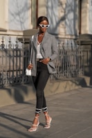 A fashionable woman walks confidently along a street. She is wearing white cat-eye sunglasses, a checkered blazer over a lace top, black skinny jeans, and white strappy high-heeled sandals. She carries a white handbag and sports a watch on her wrist. The background features an ornate wrought iron fence and a building with shadows cast on the wall.