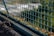A metal wire fence is depicted with a grid pattern. The fence is seen in focus, while the background appears blurred. Small green plants and moss are growing at the base of the fence, suggesting an outdoor environment, possibly along a path or garden area.