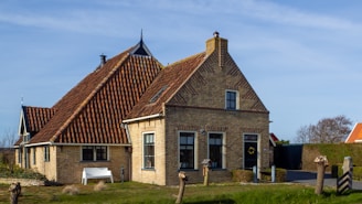 A charming house with traditional brick architecture and red-tiled roofs, situated in a rural setting. The house has multiple gables, some windows, a front door with a wreath, and a small garden area with a bench and trimmed shrubs. The lawn in the forefront is green with patches of wild grass, bordered by a low wooden fence. The sky above is clear and blue.