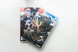 Two Nintendo Switch game cases are displayed on a light surface. The top case features 'Monster Hunter Rise' with a dynamic illustration of a fierce dragon-like creature and several warriors in action-packed poses. The case beneath it is partially obscured but shows a different monster-themed cover art.
