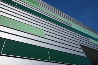 A modern building facade featuring horizontal metal panels in varying shades of green and silver. The design is geometric, with a pattern that alternates in color, giving a contemporary architectural appearance. The clear blue sky enhances the crisp lines and colors of the structure.