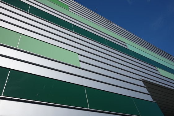 A modern building facade featuring horizontal metal panels in varying shades of green and silver. The design is geometric, with a pattern that alternates in color, giving a contemporary architectural appearance. The clear blue sky enhances the crisp lines and colors of the structure.
