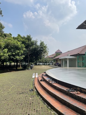 A sunny outdoor scene with a circular platform surrounded by steps. Several pairs of shoes are placed along the steps, indicating a possible entrance requirement to remove footwear. A grassy area with trees is visible to the left, providing shade. In the background, a building with a tiled roof is partially visible, adding a traditional architectural element to the scene.