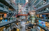 A modern shopping mall with multiple levels, featuring various stores and brand advertisements. Glass walls and ceilings allow for ample natural light. There are escalators connecting different levels and decorative elements such as statues and large hanging watches. Shoppers are seen walking around.