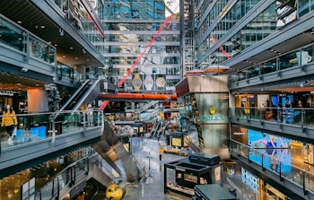 A modern shopping mall with multiple levels, featuring various stores and brand advertisements. Glass walls and ceilings allow for ample natural light. There are escalators connecting different levels and decorative elements such as statues and large hanging watches. Shoppers are seen walking around.