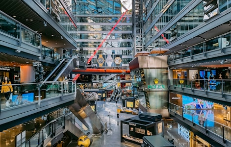 A modern shopping mall with multiple levels, featuring various stores and brand advertisements. Glass walls and ceilings allow for ample natural light. There are escalators connecting different levels and decorative elements such as statues and large hanging watches. Shoppers are seen walking around.