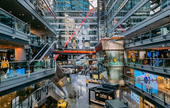 A modern shopping mall with multiple levels, featuring various stores and brand advertisements. Glass walls and ceilings allow for ample natural light. There are escalators connecting different levels and decorative elements such as statues and large hanging watches. Shoppers are seen walking around.