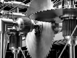 Large, interlocking metallic gears and cogs of various sizes and shapes, prominently featuring a central saw blade-like gear. The sheen of the polished metal reflects light, creating a precise, industrial aesthetic. The setup is complex and well-engineered, suggesting machinery.