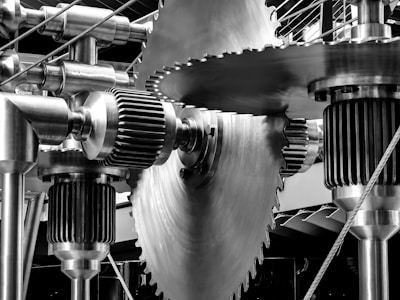 Large, interlocking metallic gears and cogs of various sizes and shapes, prominently featuring a central saw blade-like gear. The sheen of the polished metal reflects light, creating a precise, industrial aesthetic. The setup is complex and well-engineered, suggesting machinery.