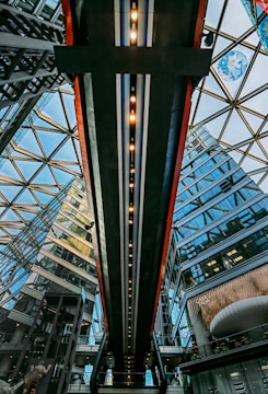 A modern architectural space with a glass roof structured in geometric patterns. The interior features a suspended walkway with evenly spaced lights. Surrounding the area are buildings with reflective glass windows, showcasing a blend of industrial and contemporary design elements.