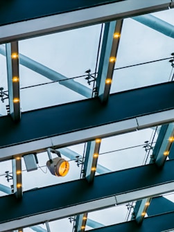 A modern ceiling structure featuring glass panels supported by metallic beams. Embedded lights illuminate the beams while a prominent spotlight is attached underneath one of the beams. The clear sky is visible through the translucent panels, creating a contrast with the industrial design.