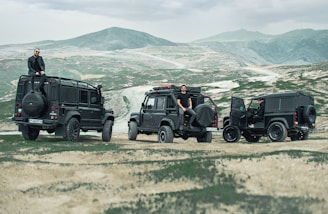A rugged outdoor setting with three black off-road vehicles parked on an arid, mountainous landscape. The vehicles are lined up, each with a person either sitting on or standing next to it. The terrain is a mix of grass and exposed earth, with distant mountains under a cloudy sky.
