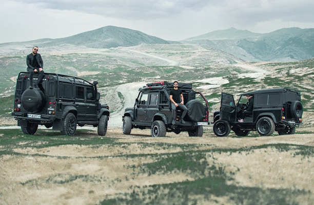 A rugged outdoor setting with three black off-road vehicles parked on an arid, mountainous landscape. The vehicles are lined up, each with a person either sitting on or standing next to it. The terrain is a mix of grass and exposed earth, with distant mountains under a cloudy sky.