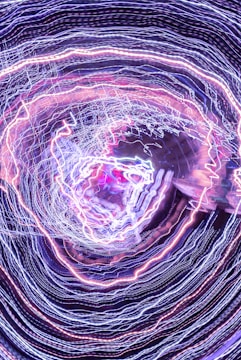 A swirling pattern of intricate light trails in various colors, primarily composed of blues, purples, and pinks. The lines create a vortex-like appearance, giving a sense of dynamic movement and energy.