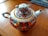 A decorative teapot adorned with intricate floral patterns featuring red, orange, and teal designs, accented with gold details. The teapot has a graceful spout and handle with gold enhancements, sitting on a wooden surface with fabric in the background.
