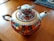 A decorative teapot adorned with intricate floral patterns featuring red, orange, and teal designs, accented with gold details. The teapot has a graceful spout and handle with gold enhancements, sitting on a wooden surface with fabric in the background.